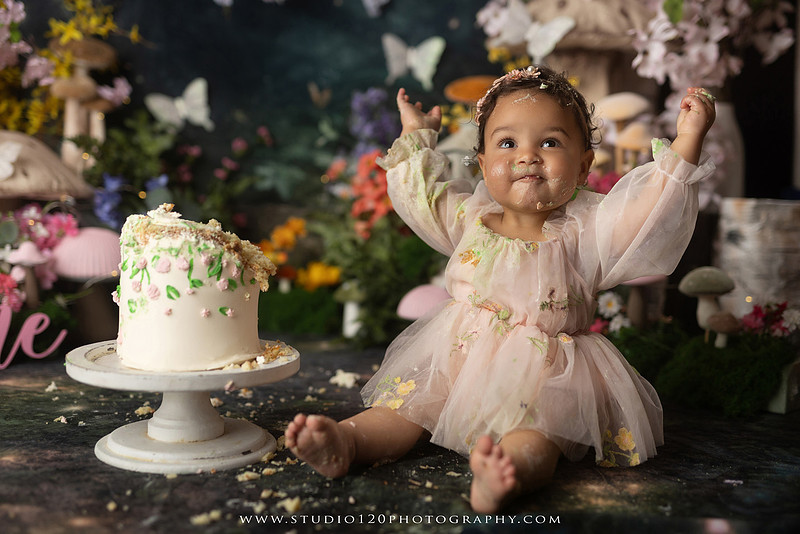 Baby girl enjoying a whimsical enchanted garden cake smash surrounded by oversized mushrooms, butterflies, blooming flowers, lush greenery, and a custom floral birthday cake while celebrating her first birthday at Studio 120 Photography in Ramona, California.