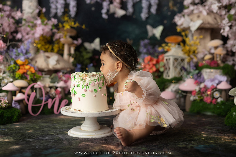 Baby girl enjoying a whimsical enchanted garden cake smash surrounded by oversized mushrooms, butterflies, blooming flowers, lush greenery, and a custom floral birthday cake while celebrating her first birthday at Studio 120 Photography in Ramona, California.