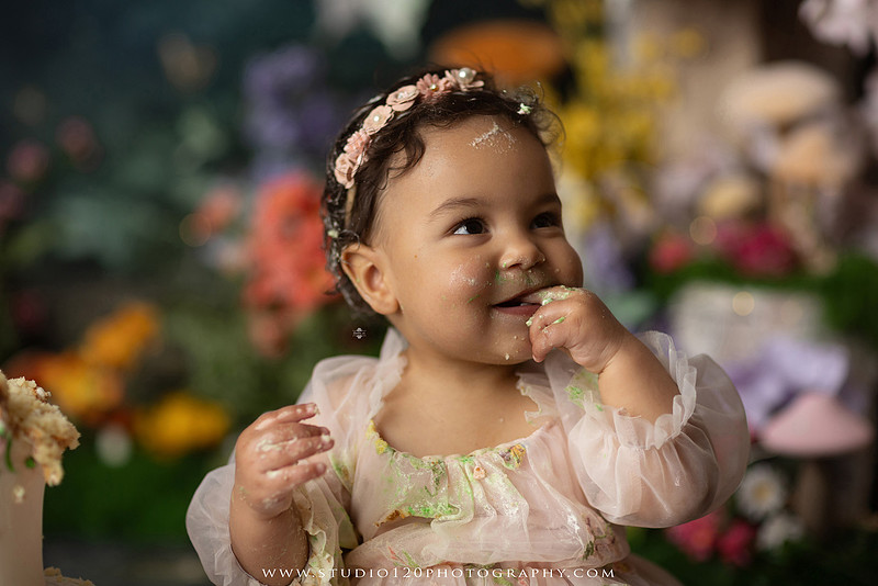 Baby girl enjoying a whimsical enchanted garden cake smash surrounded by oversized mushrooms, butterflies, blooming flowers, lush greenery, and a custom floral birthday cake while celebrating her first birthday at Studio 120 Photography in Ramona, California.