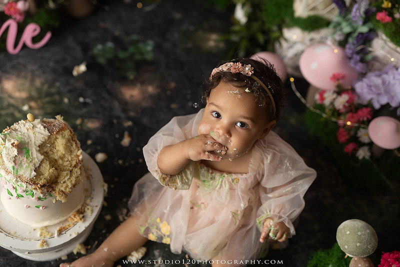 Baby girl enjoying a whimsical enchanted garden cake smash surrounded by oversized mushrooms, butterflies, blooming flowers, lush greenery, and a custom floral birthday cake while celebrating her first birthday at Studio 120 Photography in Ramona, California.
