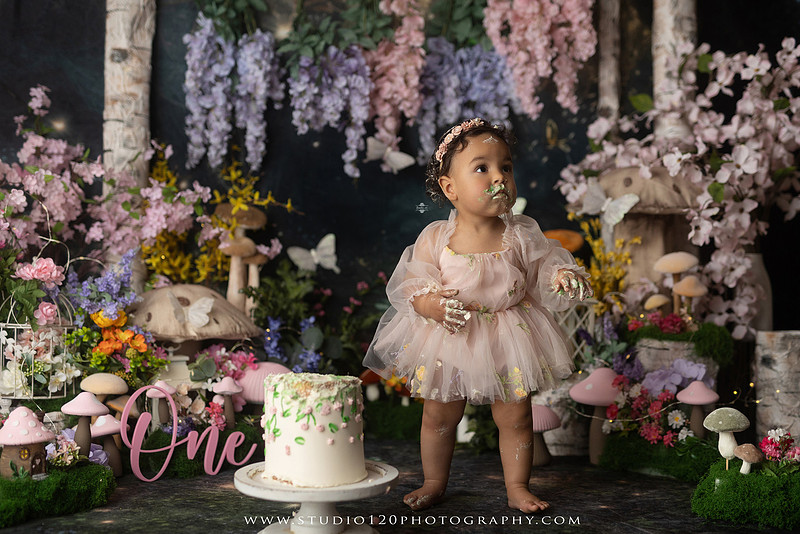 Baby girl enjoying a whimsical enchanted garden cake smash surrounded by oversized mushrooms, butterflies, blooming flowers, lush greenery, and a custom floral birthday cake while celebrating her first birthday at Studio 120 Photography in Ramona, California.
