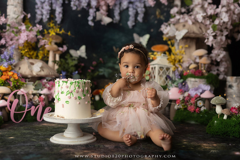 Baby girl enjoying a whimsical enchanted garden cake smash surrounded by oversized mushrooms, butterflies, blooming flowers, lush greenery, and a custom floral birthday cake while celebrating her first birthday at Studio 120 Photography in Ramona, California.