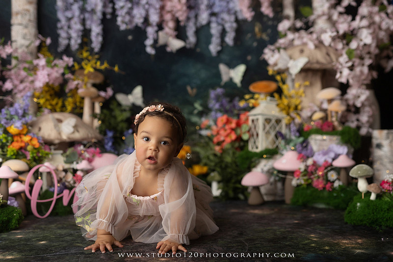 Baby girl enjoying a whimsical enchanted garden cake smash surrounded by oversized mushrooms, butterflies, blooming flowers, lush greenery, and a custom floral birthday cake while celebrating her first birthday at Studio 120 Photography in Ramona, California.