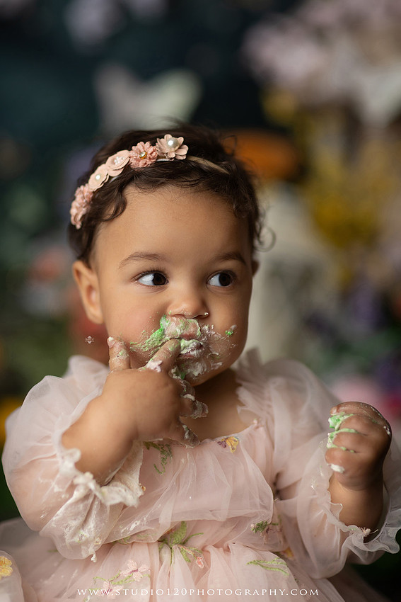 Baby girl enjoying a whimsical enchanted garden cake smash surrounded by oversized mushrooms, butterflies, blooming flowers, lush greenery, and a custom floral birthday cake while celebrating her first birthday at Studio 120 Photography in Ramona, California.