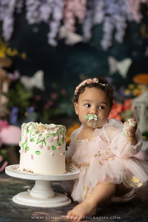 Baby girl enjoying a whimsical enchanted garden cake smash surrounded by oversized mushrooms, butterflies, blooming flowers, lush greenery, and a custom floral birthday cake while celebrating her first birthday at Studio 120 Photography in Ramona, California.