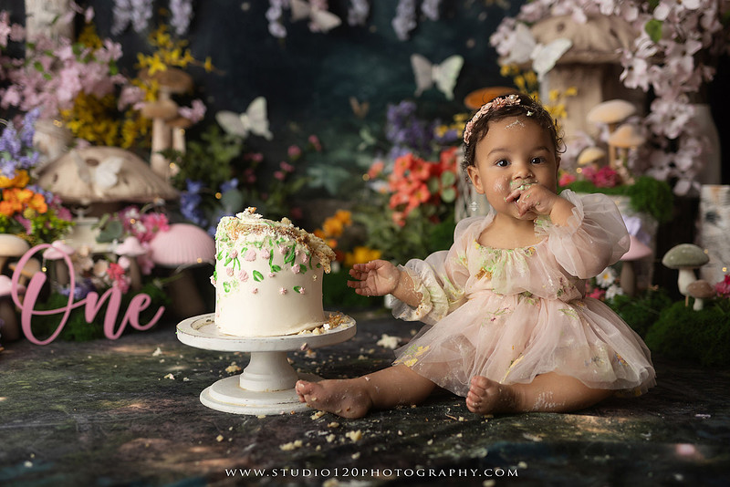 Baby girl enjoying a whimsical enchanted garden cake smash surrounded by oversized mushrooms, butterflies, blooming flowers, lush greenery, and a custom floral birthday cake while celebrating her first birthday at Studio 120 Photography in Ramona, California.