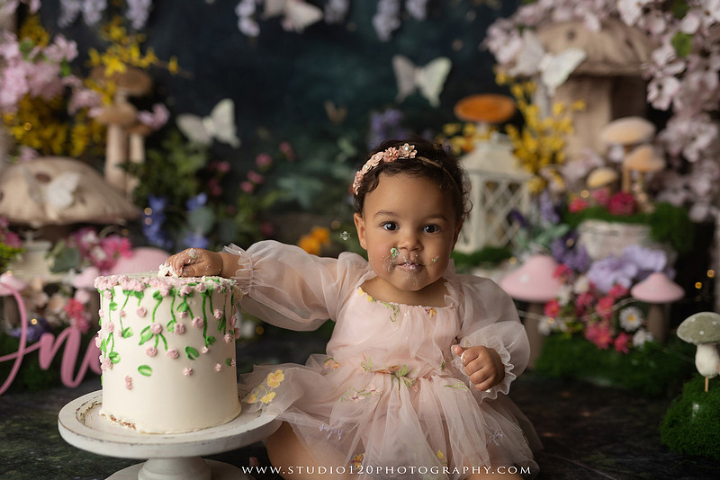 Baby girl enjoying a whimsical enchanted garden cake smash surrounded by oversized mushrooms, butterflies, blooming flowers, lush greenery, and a custom floral birthday cake while celebrating her first birthday at Studio 120 Photography in Ramona, California.