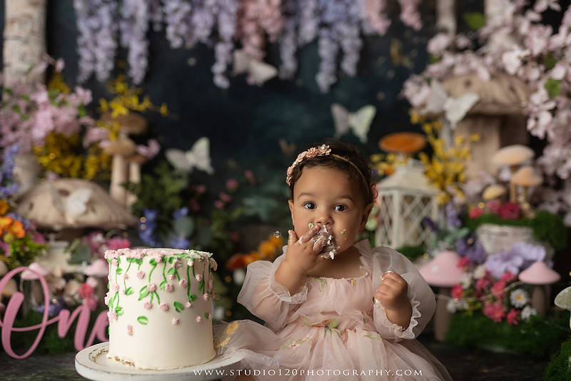Baby girl enjoying a whimsical enchanted garden cake smash surrounded by oversized mushrooms, butterflies, blooming flowers, lush greenery, and a custom floral birthday cake while celebrating her first birthday at Studio 120 Photography in Ramona, California.