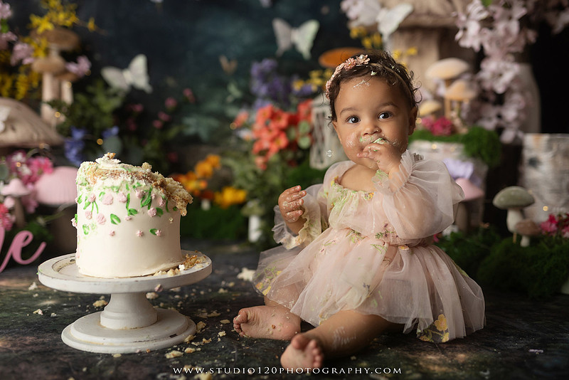 Baby girl enjoying a whimsical enchanted garden cake smash surrounded by oversized mushrooms, butterflies, blooming flowers, lush greenery, and a custom floral birthday cake while celebrating her first birthday at Studio 120 Photography in Ramona, California.