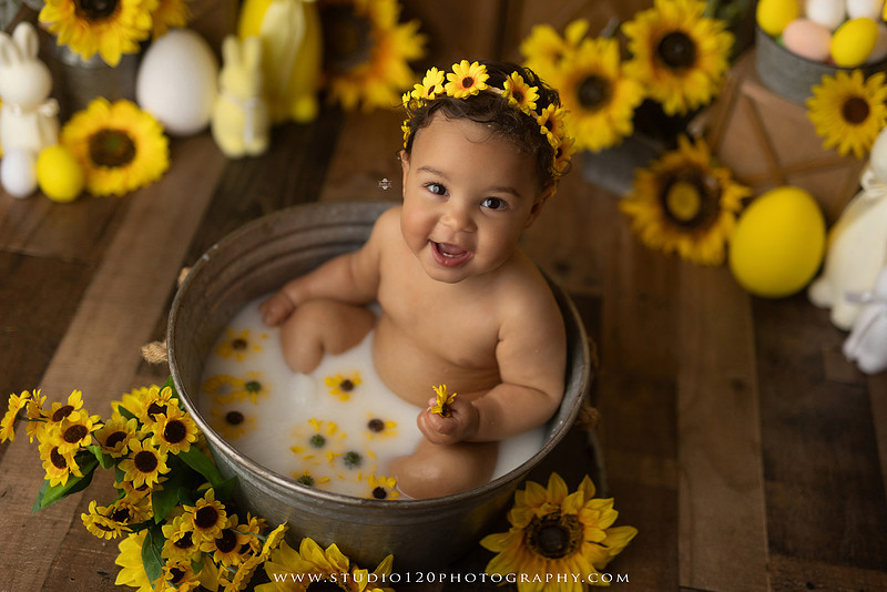 One-year-old baby girl enjoying a sunflower-themed milk bath in a vintage galvanized tub surrounded by bright yellow sunflowers, rustic spring decor, Easter accents, and cheerful floral details during a milestone photography session at Studio 120 Photography in Ramona, California.
