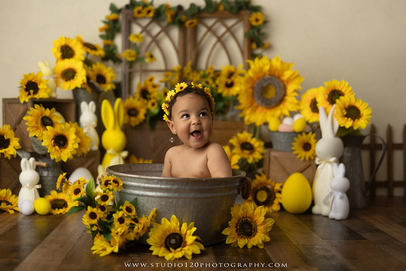 One-year-old baby girl enjoying a sunflower-themed milk bath in a vintage galvanized tub surrounded by bright yellow sunflowers, rustic spring decor, Easter accents, and cheerful floral details during a milestone photography session at Studio 120 Photography in Ramona, California.