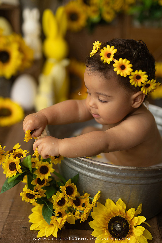 One-year-old baby girl enjoying a sunflower-themed milk bath in a vintage galvanized tub surrounded by bright yellow sunflowers, rustic spring decor, Easter accents, and cheerful floral details during a milestone photography session at Studio 120 Photography in Ramona, California.