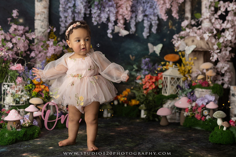 Baby girl enjoying a whimsical enchanted garden cake smash surrounded by oversized mushrooms, butterflies, blooming flowers, lush greenery, and a custom floral birthday cake while celebrating her first birthday at Studio 120 Photography in Ramona, California.