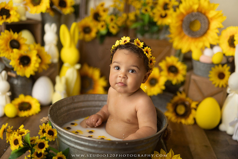 One-year-old baby girl enjoying a sunflower-themed milk bath in a vintage galvanized tub surrounded by bright yellow sunflowers, rustic spring decor, Easter accents, and cheerful floral details during a milestone photography session at Studio 120 Photography in Ramona, California.