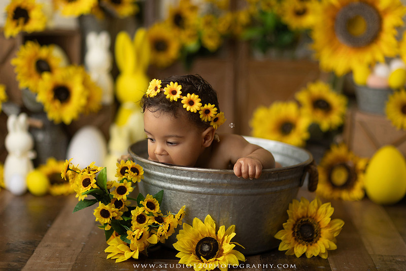 One-year-old baby girl enjoying a sunflower-themed milk bath in a vintage galvanized tub surrounded by bright yellow sunflowers, rustic spring decor, Easter accents, and cheerful floral details during a milestone photography session at Studio 120 Photography in Ramona, California.