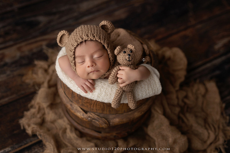 Newborn baby boy sleeping peacefully in neutral-toned wraps and rustic props, including a wooden bucket, teddy bear, and western-inspired cowboy outfit with denim overalls and a cowboy hat during a custom newborn photography session at Studio 120 Photography in Ramona, California.