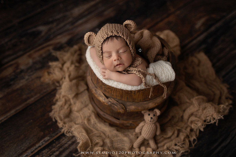 Newborn baby boy sleeping peacefully in neutral-toned wraps and rustic props, including a wooden bucket, teddy bear, and western-inspired cowboy outfit with denim overalls and a cowboy hat during a custom newborn photography session at Studio 120 Photography in Ramona, California.