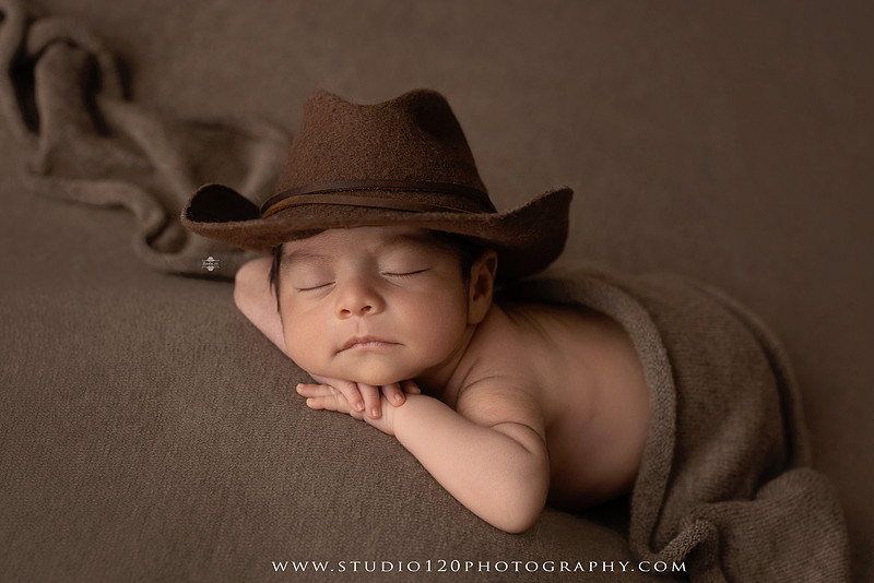 Newborn baby boy sleeping peacefully in neutral-toned wraps and rustic props, including a wooden bucket, teddy bear, and western-inspired cowboy outfit with denim overalls and a cowboy hat during a custom newborn photography session at Studio 120 Photography in Ramona, California.