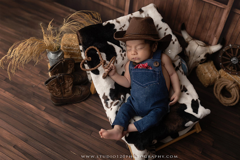 Newborn baby boy sleeping peacefully in neutral-toned wraps and rustic props, including a wooden bucket, teddy bear, and western-inspired cowboy outfit with denim overalls and a cowboy hat during a custom newborn photography session at Studio 120 Photography in Ramona, California.