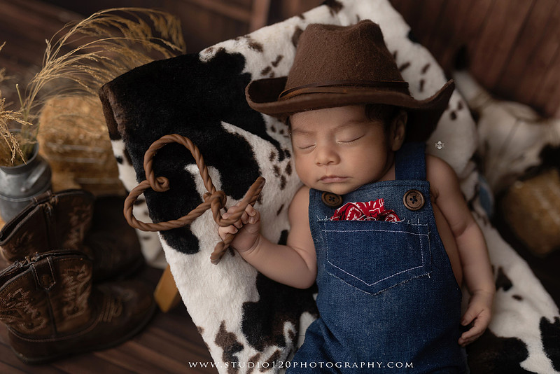 Newborn baby boy sleeping peacefully in neutral-toned wraps and rustic props, including a wooden bucket, teddy bear, and western-inspired cowboy outfit with denim overalls and a cowboy hat during a custom newborn photography session at Studio 120 Photography in Ramona, California.