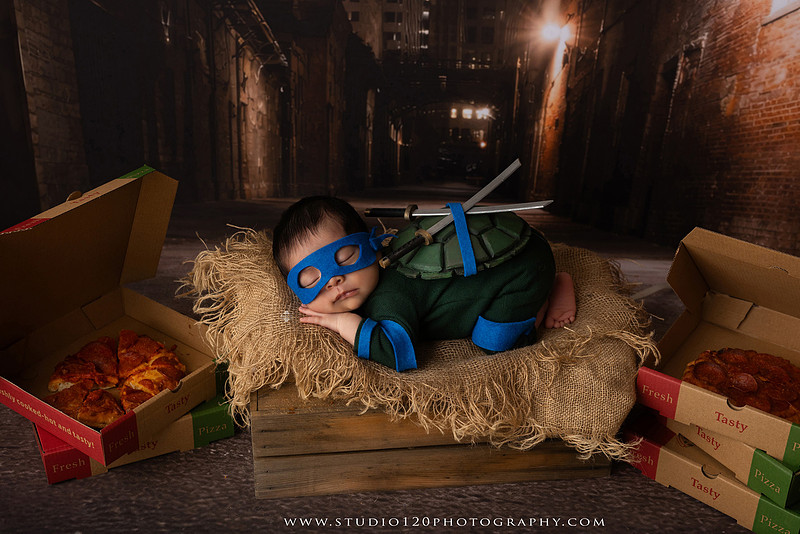 Newborn baby boy dressed in a handmade Teenage Mutant Ninja Turtles-inspired costume with a custom shell, blue mask, and 3D-printed katanas, sleeping beside pizza boxes in a creative custom newborn photography setup by Studio 120 Photography in Ramona, California.