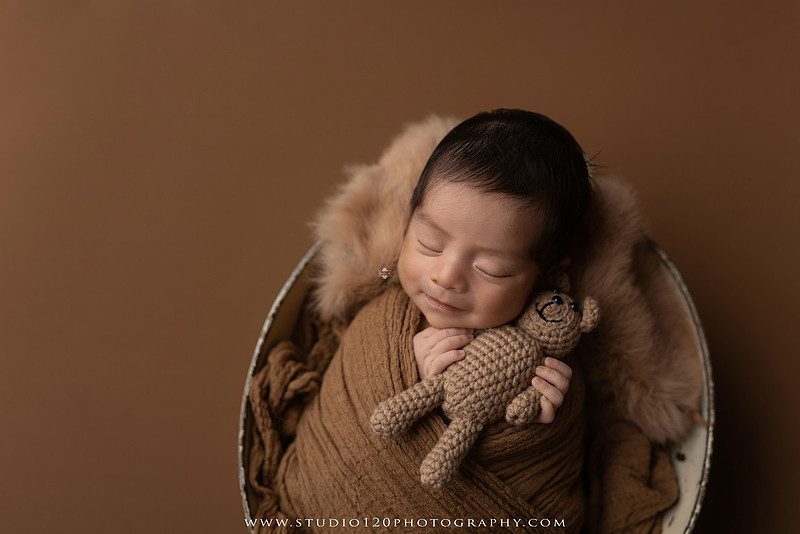 Newborn baby boy sleeping peacefully in neutral-toned wraps and rustic props, including a wooden bucket, teddy bear, and western-inspired cowboy outfit with denim overalls and a cowboy hat during a custom newborn photography session at Studio 120 Photography in Ramona, California.