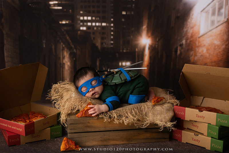 Newborn baby boy dressed in a handmade Teenage Mutant Ninja Turtles-inspired costume with a custom shell, blue mask, and 3D-printed katanas, sleeping beside pizza boxes in a creative custom newborn photography setup by Studio 120 Photography in Ramona, California.
