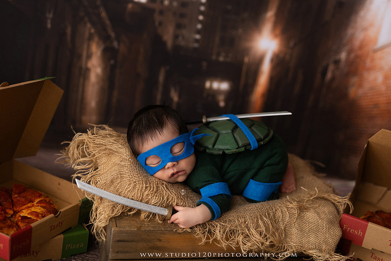 Newborn baby boy dressed in a handmade Teenage Mutant Ninja Turtles-inspired costume with a custom shell, blue mask, and 3D-printed katanas, sleeping beside pizza boxes in a creative custom newborn photography setup by Studio 120 Photography in Ramona, California.