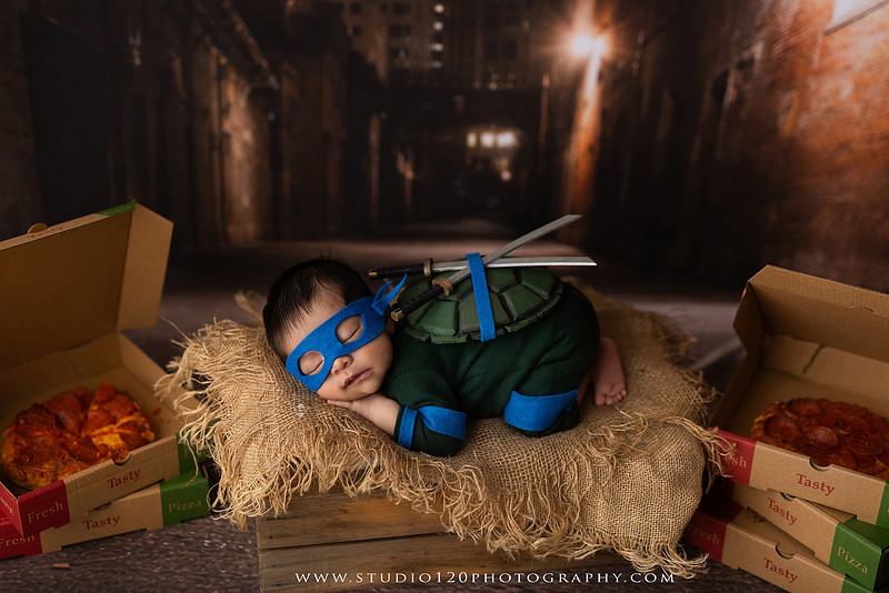 Newborn baby boy dressed in a handmade Teenage Mutant Ninja Turtles-inspired costume with a custom shell, blue mask, and 3D-printed katanas, sleeping beside pizza boxes in a creative custom newborn photography setup by Studio 120 Photography in Ramona, California.