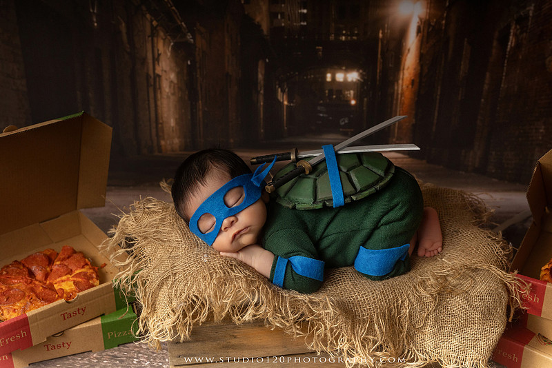 Newborn baby boy dressed in a handmade Teenage Mutant Ninja Turtles-inspired costume with a custom shell, blue mask, and 3D-printed katanas, sleeping beside pizza boxes in a creative custom newborn photography setup by Studio 120 Photography in Ramona, California.