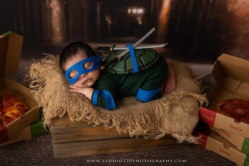 Newborn baby boy dressed in a handmade Teenage Mutant Ninja Turtles-inspired costume with a custom shell, blue mask, and 3D-printed katanas, sleeping beside pizza boxes in a creative custom newborn photography setup by Studio 120 Photography in Ramona, California.