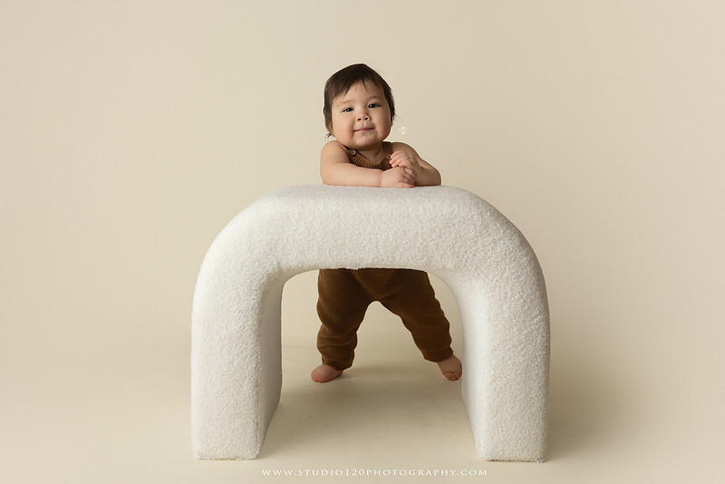 Smiling one-year-old boy in a knit romper sitting on a wooden stool during a milestone portrait session at Studio 120 Photography in Ramona, California.