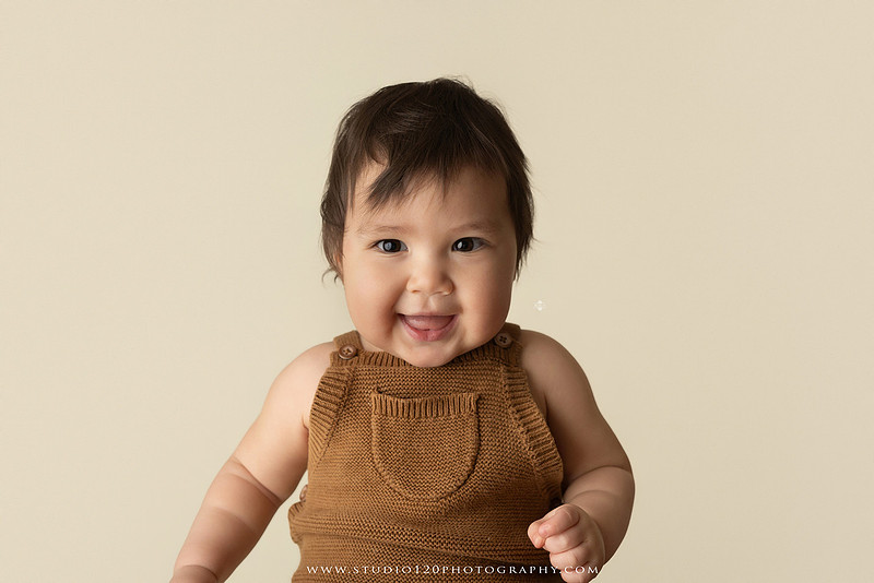 Smiling one-year-old boy in a knit romper sitting on a wooden stool during a milestone portrait session at Studio 120 Photography in Ramona, California.
