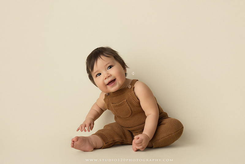 Smiling one-year-old boy in a knit romper sitting on a wooden stool during a milestone portrait session at Studio 120 Photography in Ramona, California.