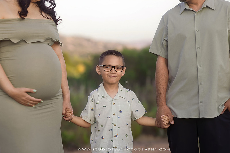 A golden hour maternity photography session at Lake Poway featuring an expecting family surrounded by wildflowers, rolling hills, and mountain views. The gallery includes romantic portraits of the parents, family photos with their young son, candid moments celebrating a growing family, and close-up images highlighting the mother's baby bump and the excitement of becoming a big brother. Captured in soft natural light with timeless neutral tones by Studio 120 Photography in Ramona, serving families throughout San Diego.