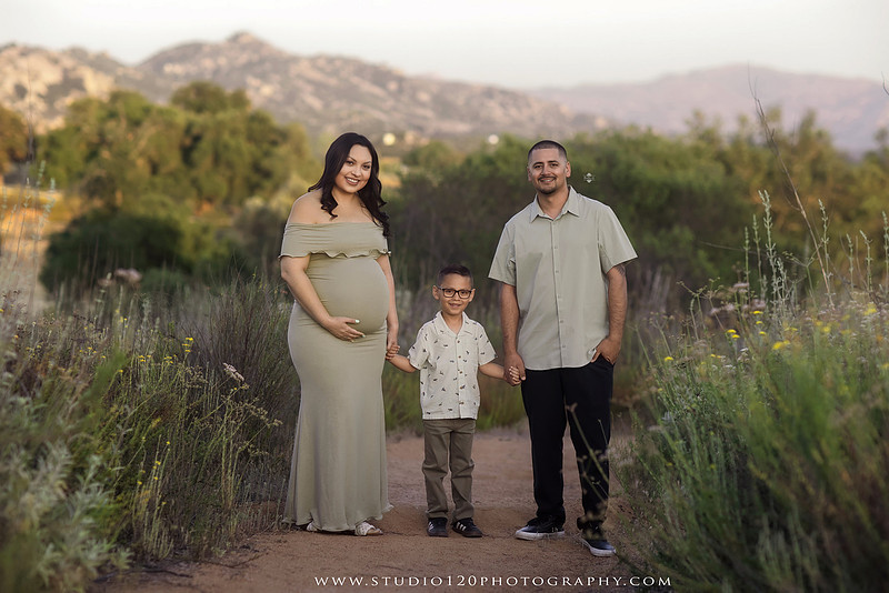 A golden hour maternity photography session at Lake Poway featuring an expecting family surrounded by wildflowers, rolling hills, and mountain views. The gallery includes romantic portraits of the parents, family photos with their young son, candid moments celebrating a growing family, and close-up images highlighting the mother's baby bump and the excitement of becoming a big brother. Captured in soft natural light with timeless neutral tones by Studio 120 Photography in Ramona, serving families throughout San Diego.