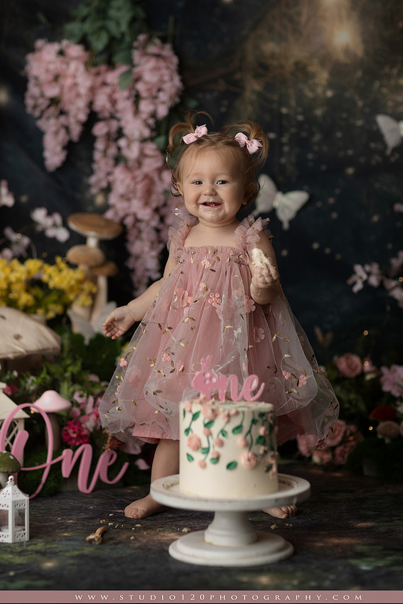 One-year-old girl celebrating her first birthday with an enchanted fairy garden cake smash, wearing a blush pink fairy dress and wings while exploring a whimsical set filled with flowers, butterflies, woodland mushrooms, lanterns, and a floral cake. Custom fairy-themed first birthday photography by Studio 120 Photography in Ramona, serving San Diego and surrounding areas.