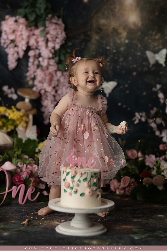 One-year-old girl celebrating her first birthday with an enchanted fairy garden cake smash, wearing a blush pink fairy dress and wings while exploring a whimsical set filled with flowers, butterflies, woodland mushrooms, lanterns, and a floral cake. Custom fairy-themed first birthday photography by Studio 120 Photography in Ramona, serving San Diego and surrounding areas.