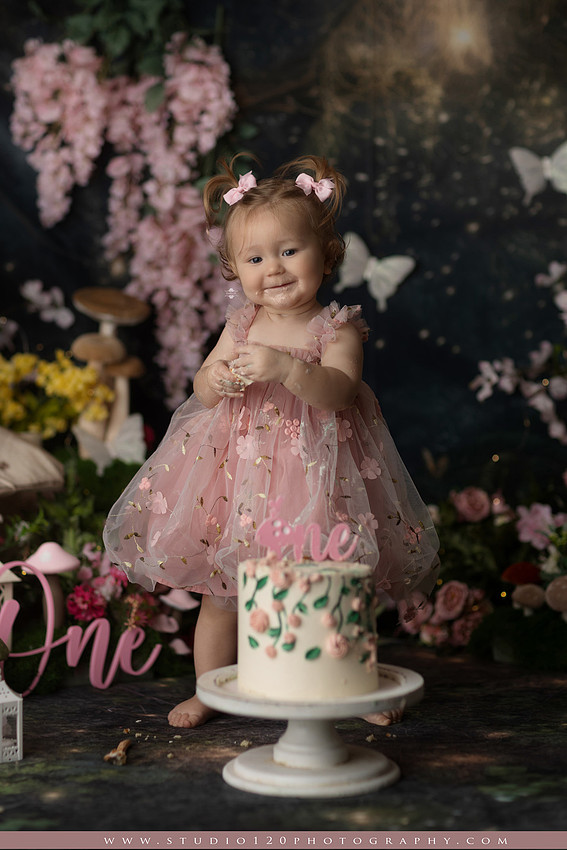 One-year-old girl celebrating her first birthday with an enchanted fairy garden cake smash, wearing a blush pink fairy dress and wings while exploring a whimsical set filled with flowers, butterflies, woodland mushrooms, lanterns, and a floral cake. Custom fairy-themed first birthday photography by Studio 120 Photography in Ramona, serving San Diego and surrounding areas.