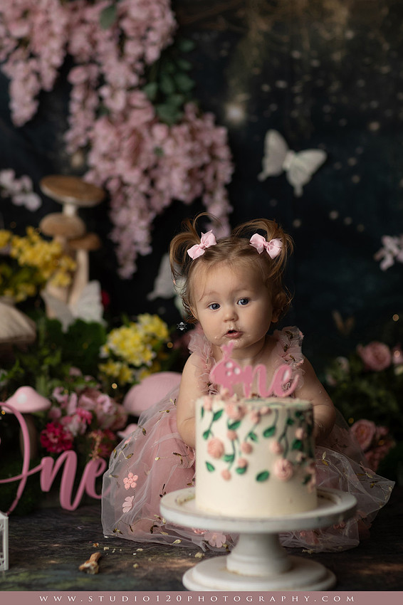 One-year-old girl celebrating her first birthday with an enchanted fairy garden cake smash, wearing a blush pink fairy dress and wings while exploring a whimsical set filled with flowers, butterflies, woodland mushrooms, lanterns, and a floral cake. Custom fairy-themed first birthday photography by Studio 120 Photography in Ramona, serving San Diego and surrounding areas.