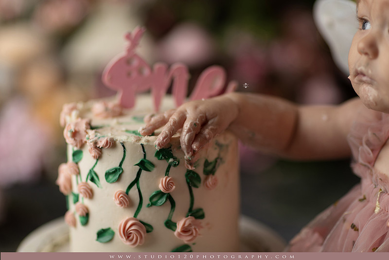 One-year-old girl celebrating her first birthday with an enchanted fairy garden cake smash, wearing a blush pink fairy dress and wings while exploring a whimsical set filled with flowers, butterflies, woodland mushrooms, lanterns, and a floral cake. Custom fairy-themed first birthday photography by Studio 120 Photography in Ramona, serving San Diego and surrounding areas.