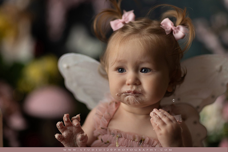 One-year-old girl celebrating her first birthday with an enchanted fairy garden cake smash, wearing a blush pink fairy dress and wings while exploring a whimsical set filled with flowers, butterflies, woodland mushrooms, lanterns, and a floral cake. Custom fairy-themed first birthday photography by Studio 120 Photography in Ramona, serving San Diego and surrounding areas.