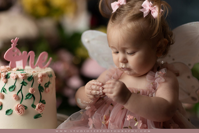 One-year-old girl celebrating her first birthday with an enchanted fairy garden cake smash, wearing a blush pink fairy dress and wings while exploring a whimsical set filled with flowers, butterflies, woodland mushrooms, lanterns, and a floral cake. Custom fairy-themed first birthday photography by Studio 120 Photography in Ramona, serving San Diego and surrounding areas.