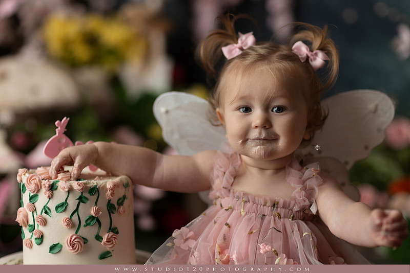 One-year-old girl celebrating her first birthday with an enchanted fairy garden cake smash, wearing a blush pink fairy dress and wings while exploring a whimsical set filled with flowers, butterflies, woodland mushrooms, lanterns, and a floral cake. Custom fairy-themed first birthday photography by Studio 120 Photography in Ramona, serving San Diego and surrounding areas.