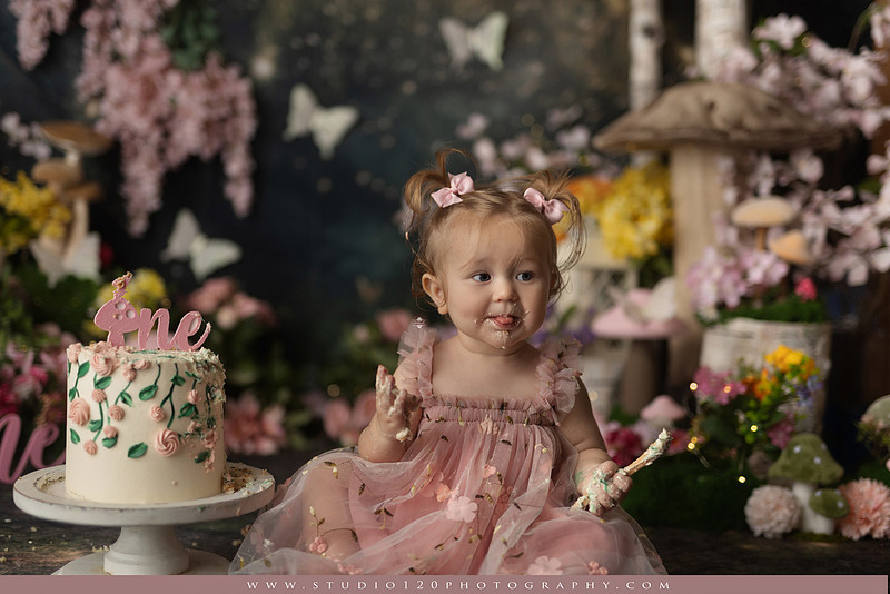 One-year-old girl celebrating her first birthday with an enchanted fairy garden cake smash, wearing a blush pink fairy dress and wings while exploring a whimsical set filled with flowers, butterflies, woodland mushrooms, lanterns, and a floral cake. Custom fairy-themed first birthday photography by Studio 120 Photography in Ramona, serving San Diego and surrounding areas.