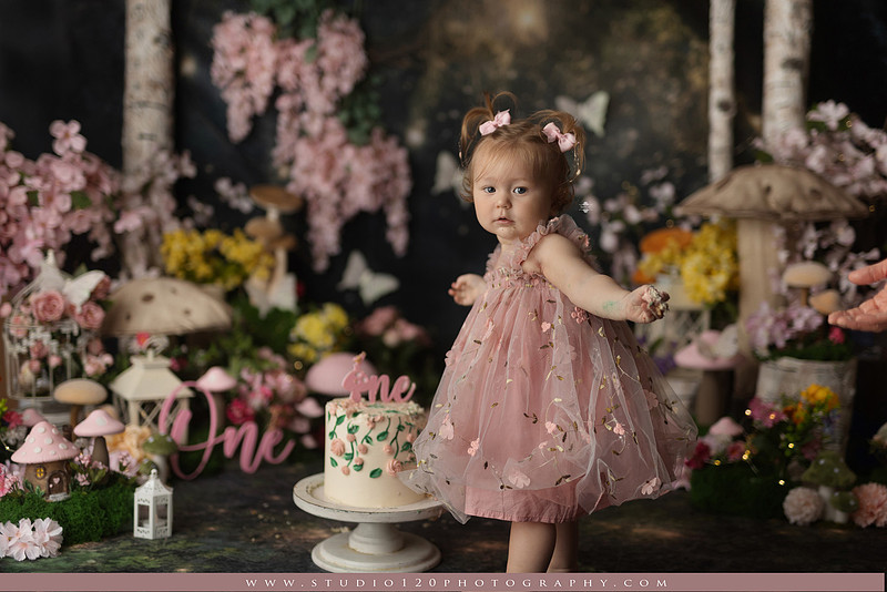 One-year-old girl celebrating her first birthday with an enchanted fairy garden cake smash, wearing a blush pink fairy dress and wings while exploring a whimsical set filled with flowers, butterflies, woodland mushrooms, lanterns, and a floral cake. Custom fairy-themed first birthday photography by Studio 120 Photography in Ramona, serving San Diego and surrounding areas.
