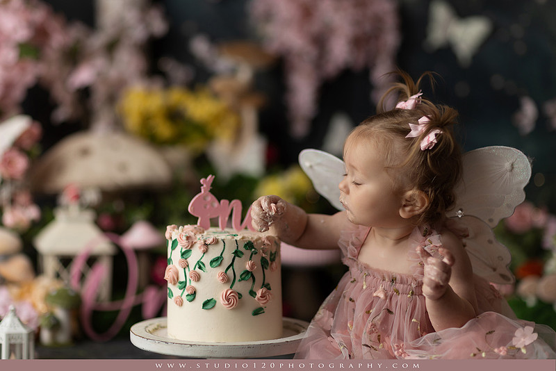 One-year-old girl celebrating her first birthday with an enchanted fairy garden cake smash, wearing a blush pink fairy dress and wings while exploring a whimsical set filled with flowers, butterflies, woodland mushrooms, lanterns, and a floral cake. Custom fairy-themed first birthday photography by Studio 120 Photography in Ramona, serving San Diego and surrounding areas.