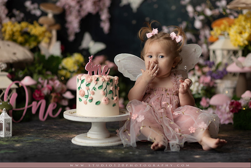 One-year-old girl celebrating her first birthday with an enchanted fairy garden cake smash, wearing a blush pink fairy dress and wings while exploring a whimsical set filled with flowers, butterflies, woodland mushrooms, lanterns, and a floral cake. Custom fairy-themed first birthday photography by Studio 120 Photography in Ramona, serving San Diego and surrounding areas.