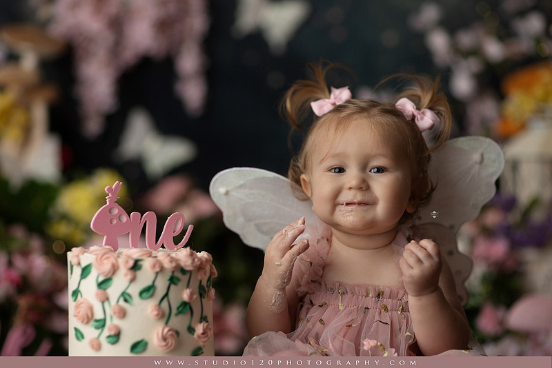One-year-old girl celebrating her first birthday with an enchanted fairy garden cake smash, wearing a blush pink fairy dress and wings while exploring a whimsical set filled with flowers, butterflies, woodland mushrooms, lanterns, and a floral cake. Custom fairy-themed first birthday photography by Studio 120 Photography in Ramona, serving San Diego and surrounding areas.