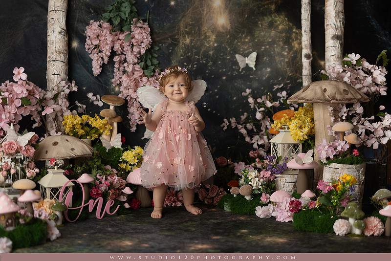 One-year-old girl celebrating her first birthday with an enchanted fairy garden cake smash, wearing a blush pink fairy dress and wings while exploring a whimsical set filled with flowers, butterflies, woodland mushrooms, lanterns, and a floral cake. Custom fairy-themed first birthday photography by Studio 120 Photography in Ramona, serving San Diego and surrounding areas.